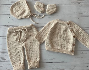 May include: A knitted baby outfit set in a cream color. The set includes a sweater, pants, a hat, and two pairs of booties.