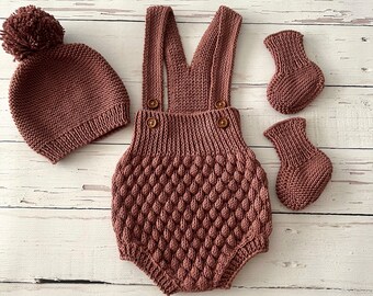 Könnte beinhalten: Ein gestricktes Baby-Outfit-Set in staubigem Rosaton. Das Set beinhaltet eine Mütze mit Bommel, ein Paar Booties und einen einteiligen Strampler mit Trägern.