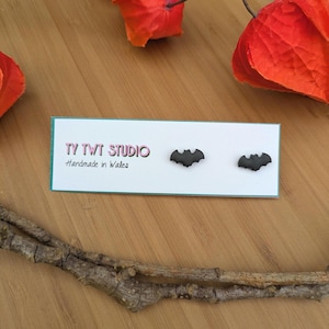 May include: Black bat-shaped earrings on a white card with the text "TY TWT STUDIO Handmade in Wales". The earrings are displayed on a wooden surface, with orange floral accents and a twig.