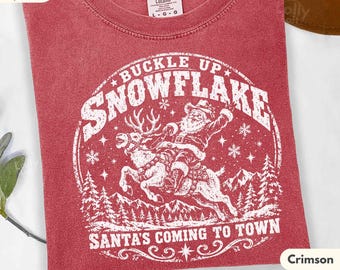 May include: A crimson red t-shirt featuring a white graphic print of Santa Claus riding a reindeer. The text reads BUCKLE UP SNOWFLAKE and SANTA'S COMING TO TOWN. The design includes snowflakes, pine trees, and mountain silhouettes in a circular frame.