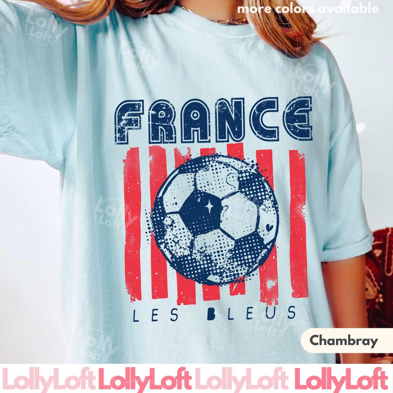 May include: Light blue t-shirt with a distressed graphic featuring the word "FRANCE" above a soccer ball and red stripes. The text "LES BLEUS" is below the ball. The shirt is labeled "Chambray".