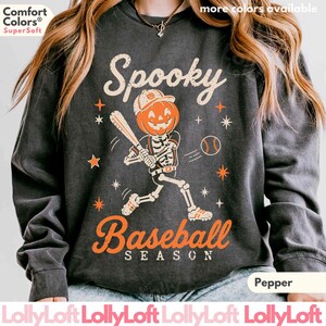 May include: A charcoal gray crewneck sweatshirt featuring a graphic of a skeleton with a pumpkin head playing baseball. The text reads Spooky Baseball Season in orange and cream. The design includes small orange stars and a baseball.