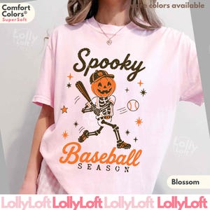 May include: A light pink short-sleeve t-shirt featuring a graphic of a skeleton with a pumpkin head wearing a baseball cap and holding a bat. The text reads Spooky Baseball Season, surrounded by small orange and black star and baseball icons.