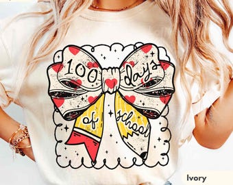 May include: An ivory t-shirt featuring a large graphic of a bow made from yellow pencils with red erasers. The text 100 days of school is written in black script across the bow, which is decorated with small red hearts and black stars.