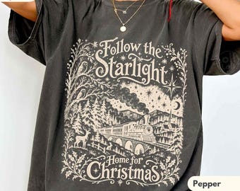 May include: A charcoal grey short-sleeve t-shirt featuring a cream-coloured graphic print of a steam train crossing a bridge, a deer, trees, and stars. The text reads Follow the Starlight Home for Christmas in a decorative font style.