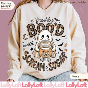 May include: An ivory crewneck sweatshirt featuring a graphic of a ghost holding a coffee cup with a jack-o'-lantern face. The text reads "freshly BOO'D with SCREAM & SUGAR" and "SPOOKY BREW" on the cup, surrounded by small bats, stars, and crescent moons.