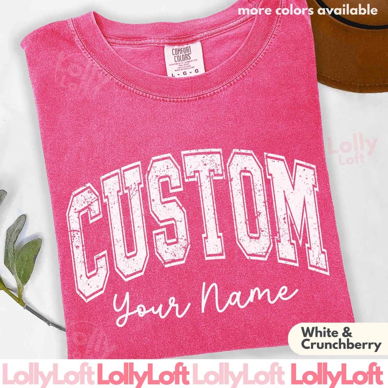 以下が含まれることがあります： 白のブロック体で「CUSTOM」、筆記体で「your Name」と書かれたピンク色のTシャツ。Comfort Colorsブランドで、色は「White & Crunchberry」です。クルーネックで半袖のTシャツです。