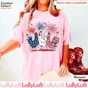 Pode incluir: Uma camiseta rosa com um gr&aacute;fico de tr&ecirc;s galinhas. As galinhas usam cores vermelho, branco e azul, que lembram a bandeira portuguesa. O gr&aacute;fico tamb&eacute;m inclui fogos de artif&iacute;cio no fundo. O texto "Comfort Colors&reg; Super Soft" &eacute; vis&iacute;vel no canto superior esquerdo da imagem. O texto "Mais cores dispon&iacute;veis" &eacute; vis&iacute;vel no canto superior direito da imagem.