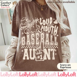 May include: A brown short-sleeve t-shirt featuring a cream-colored graphic with a baseball, a megaphone, and the text "Somebody's LOUD MOUTH BASEBALL AUNT #1". The shirt has a crew neckline and a relaxed fit, with a vintage-inspired distressed print design.