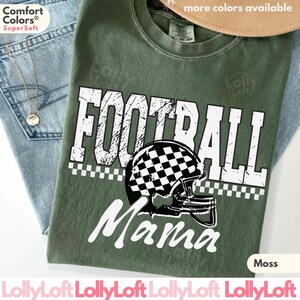 May include: Moss green t-shirt with the words "FOOTBALL Mama" in white, featuring a checkered football helmet graphic. The shirt is made of a soft material and is displayed with blue jeans and a necklace.