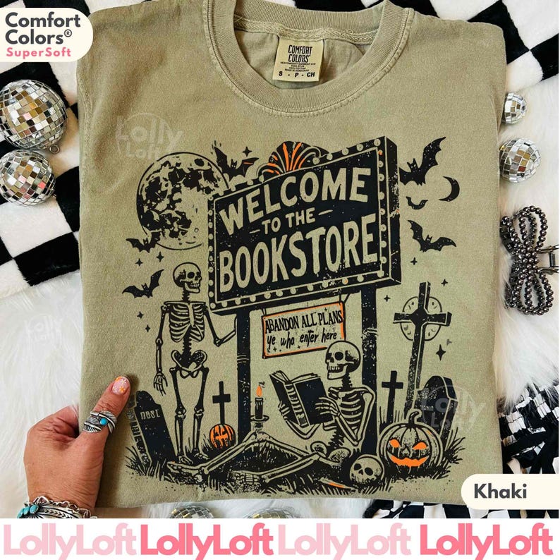 May include: Khaki-colored t-shirt with a Halloween-themed graphic. The design features skeletons, bats, pumpkins, and a sign that reads "WELCOME TO THE BOOKSTORE." The shirt is made of Comfort Colors SuperSoft material.