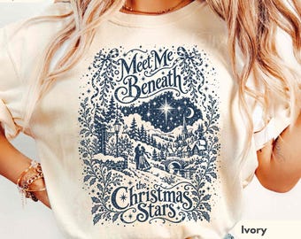 May include: An ivory short-sleeve t-shirt featuring a dark blue graphic print of a snowy winter village scene with a couple, a bridge, and a star. The text reads Meet Me Beneath the Christmas Stars in a decorative script font.
