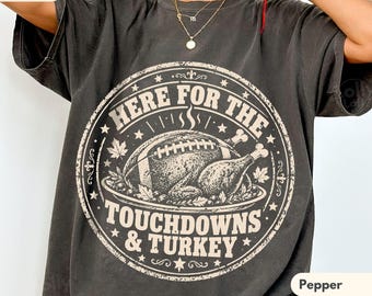May include: A charcoal grey short-sleeve t-shirt featuring a circular graphic with a roasted turkey and a football. The text reads HERE FOR THE TOUCHDOWNS & TURKEY. The design includes stars and maple leaves in a distressed, off-white colour.