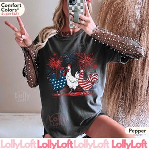 Pode incluir: Uma camiseta cinza escura com um gr&aacute;fico de tr&ecirc;s galos com cores patri&oacute;ticas vermelho, branco e azul. Os gallos est&atilde;o em p&eacute; em frente a um show de fogos de artif&iacute;cio. A camiseta tem o texto "Comfort Colors&reg; Super Soft" na parte superior.