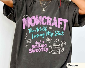 May include: A charcoal grey short-sleeve t-shirt featuring the pink text MOMCRAFT, the teal text The Art Of Losing My Shit, and the pink text And Smiling SWEETLY, accompanied by a graphic of a coffee mug and a small skull icon.