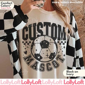 May include: An ivory t-shirt featuring a black graphic of a soccer ball centered between the words CUSTOM and MASCOT in a bold, distressed font. The shirt is worn under a black and white checkered long-sleeve garment with a Comfort Colors label.