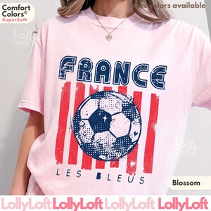 May include: A light pink t-shirt with a distressed graphic featuring the word "FRANCE" in blue, red stripes, and a soccer ball. The text "LES BLEUS" is below the ball. The shirt is labeled "Blossom" and "Comfort Colors Super Soft".