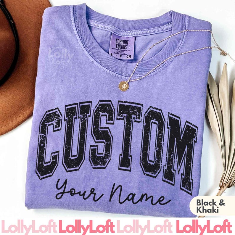 以下が含まれることがあります： 大きな黒い文字で「CUSTOM」と書かれた、色あせたライラック色のTシャツ。その下に「Your Name」という文字が筆記体で書かれています。金色のネックレスとペンダントがシャツの上にあります。シャツにはComfort Colorsのラベルが付いています。