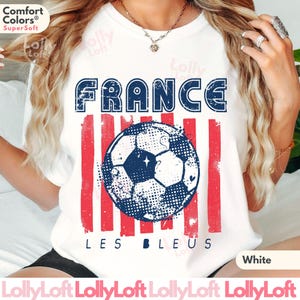 May include: White t-shirt with a distressed design featuring the word "FRANCE" in blue, red stripes, and a soccer ball graphic. The text "LES BLEUS" is below the soccer ball. The shirt is made of a soft material.