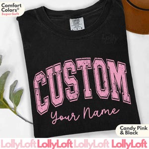 以下が含まれることがあります： 前面にピンク色の「CUSTOM」の文字、下に筆記体で「Your Name」と書かれた黒いTシャツ。「Comfort Colors」と「Super Soft」のラベルが付いています。「Candy Pink & Black」の文字も表示されています。