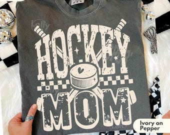 May include: A dark gray t-shirt with the words "HOCKEY MOM" in a distressed white font. The design includes hockey sticks and a puck. The shirt is labeled "Comfort Colors" and "Super Soft."