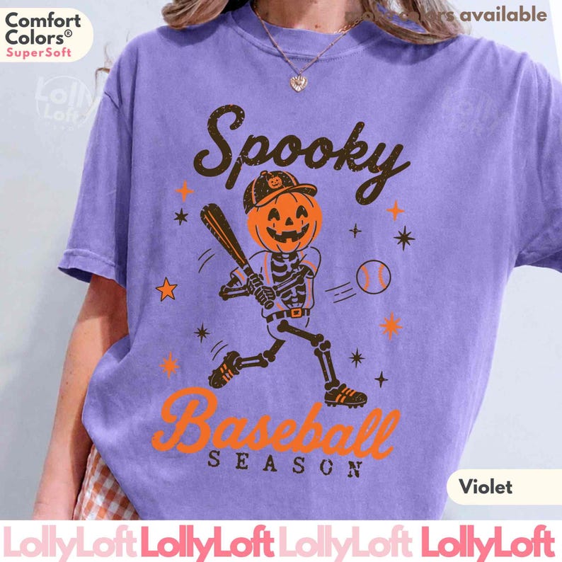 May include: A violet short-sleeve t-shirt featuring a graphic of a skeleton with a jack-o'-lantern head playing baseball. The text reads Spooky Baseball Season in black and orange. The design includes orange stars and a baseball.