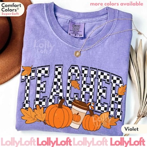 May include: A violet Comfort Colors t-shirt with the word "TEACHER" in a checkered pattern, pumpkins, and fall leaves. A gold necklace and a brown hat are visible. The shirt has the text "more colors available" and "Violet".