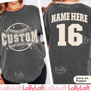 May include: A charcoal gray short-sleeve t-shirt featuring a baseball graphic with the word CUSTOM on the front. The back displays the text NAME HERE above the number 16 in large cream-colored lettering. The shirt has a crew neckline.