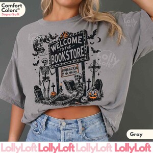 May include: Gray Comfort Colors t-shirt with a Halloween-themed bookstore graphic. The design features skeletons, bats, a full moon, tombstones, and a sign that reads "WELCOME TO THE BOOKSTORE". The text "ABANDON ALL PLANS ye who enter here" is also included.