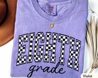 Puede incluir: Una camiseta de color violeta con las palabras "EIGHTH grade" en un diseño a cuadros blanco y negro. La camiseta tiene cuello redondo y una pequeña etiqueta que dice "COMFORT COLORS". Un collar dorado con un colgante está sobre la camiseta.