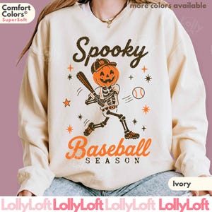 May include: A cream-colored crewneck sweatshirt featuring a graphic of a skeleton with a pumpkin head playing baseball. The text reads Spooky Baseball Season in black and orange. The design includes small orange and black stars and a baseball.