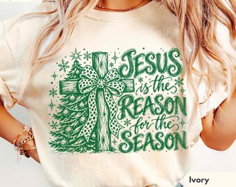 May include: An ivory t-shirt featuring a green graphic print of a Christmas tree and a cross tied with a leopard print bow. The text reads Jesus is the reason for the season. The shirt includes Comfort Colors branding and ivory colour label.