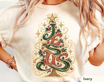 May include: An ivory t-shirt featuring a graphic of a Christmas tree formed by the word JESUS in stylised green and red lettering. The design includes gold stars, red ornaments, and green pine branches. The text Comfort Colors SuperSoft is visible.