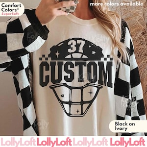 May include: An ivory crewneck sweatshirt featuring a black graphic print of a baseball catcher mask with the number 37 and the word CUSTOM. The sweatshirt is worn with a black and white checkered long-sleeve shirt layered over the top.
