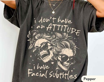 May include: A charcoal grey short-sleeve t-shirt featuring a graphic of two skulls wearing sunglasses with messy buns. The text reads, I don't have an ATTITUDE i have Facial Subtitles. The design is printed in a light cream colour on the front.