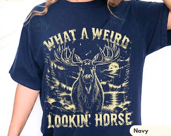 May include: A navy blue short-sleeve t-shirt featuring a gold graphic of a moose standing in a forest by a lake. The text "WHAT A WEIRD LOOKIN' HORSE" is printed in gold above and below the moose illustration. The shirt has a crew neckline.