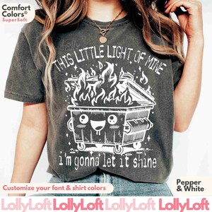 Pu&ograve; includere: Una maglietta grigia con una grafica bianca di un cassonetto dei rifiuti di cartone in fiamme con il testo "This little light of mine I'm gonna let it shine".