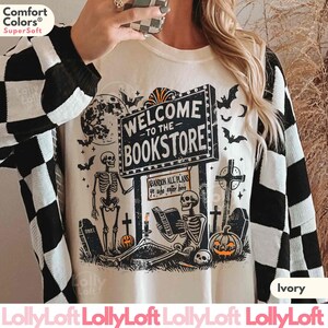 May include: Ivory-colored sweatshirt with a Halloween-themed graphic. The design features skeletons, bats, tombstones, and a sign that reads "Welcome to the Bookstore." The sweatshirt has a black and white checkered pattern on the sleeves.