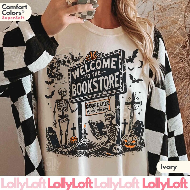May include: Ivory-colored sweatshirt with a Halloween-themed graphic. The design features skeletons, bats, tombstones, and a sign that reads "Welcome to the Bookstore." The sweatshirt has a black and white checkered pattern on the sleeves.