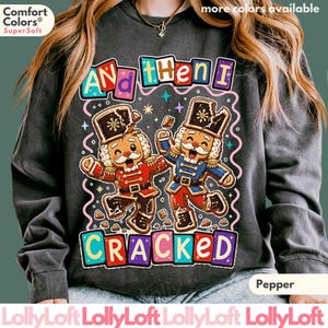 May include: A charcoal gray crewneck sweatshirt featuring a graphic of two gingerbread nutcrackers with cracked heads. The text reads "AND THEN I CRACKED" in colorful block letters. The design includes small stars and pink squiggly lines.