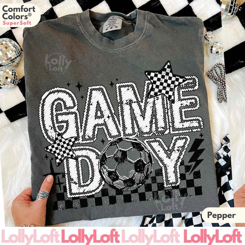 May include: Pepper-colored t-shirt with the words "GAME DAY" in white, featuring a soccer ball and checkered stars. The shirt is made of Comfort Colors SuperSoft material.