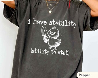 May include: A charcoal gray short-sleeve t-shirt featuring a white graphic of a small bird holding a knife. The text reads i have stability (ability to stab) in a white font. The shirt has a crew neckline and a relaxed, loose-fitting silhouette.