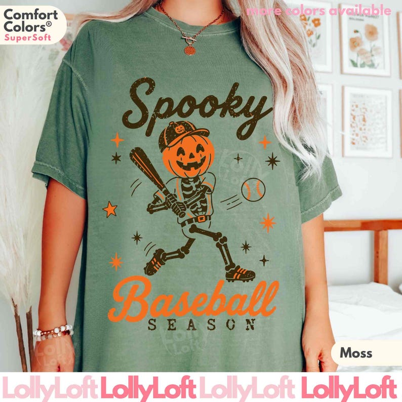 May include: A sage green short-sleeve t-shirt featuring a graphic of a skeleton with a pumpkin head playing baseball. The text reads Spooky Baseball Season in dark brown and orange. The shirt includes a Comfort Colors logo on the upper left.