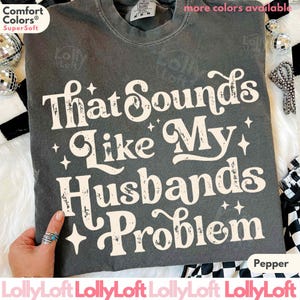 May include: Pepper-colored t-shirt with the phrase "That Sounds Like My Husbands Problem" in a distressed white font. The shirt is a Comfort Colors Super Soft tee. Additional text includes "more colors available".