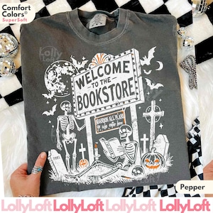 May include: Pepper-colored t-shirt with a spooky bookstore design. The graphic includes a sign that reads "Welcome to the Bookstore," skeletons, bats, a pumpkin, and tombstones. The shirt is made of Comfort Colors Super Soft material.