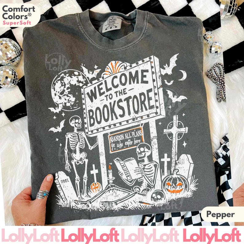 May include: Pepper-colored t-shirt with a spooky bookstore design. The graphic includes a sign that reads "Welcome to the Bookstore," skeletons, bats, a pumpkin, and tombstones. The shirt is made of Comfort Colors Super Soft material.