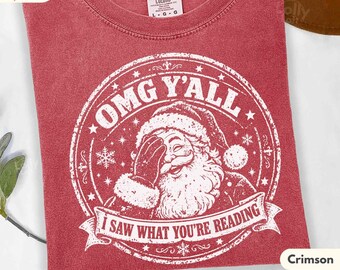 May include: A crimson t-shirt featuring a white graphic of Santa Claus with a hand over his face. The circular design includes the text OMG Y'ALL and I SAW WHAT YOU'RE READING, surrounded by stars and snowflakes. The shirt has a Comfort Colors label.
