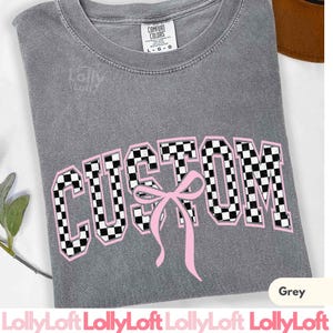 May include: A grey t-shirt with the word "CUSTOM" in a checkered pattern with a pink bow. The shirt has a crew neck and short sleeves. The brand name "Comfort Colors" is visible on the tag.