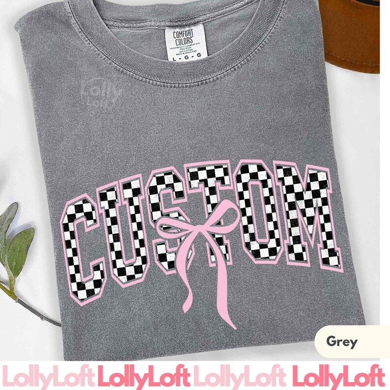 May include: A grey t-shirt with the word "CUSTOM" in a checkered pattern with a pink bow. The shirt has a crew neck and short sleeves. The brand name "Comfort Colors" is visible on the tag.