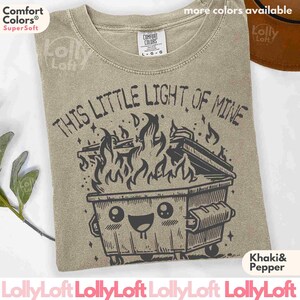 Pu&ograve; includere: Maglietta color cachi con un'illustrazione in bianco e nero di un cassonetto della spazzatura in fiamme con un viso sorridente. Il testo "This Little Light of Mine" &egrave; sopra il cassonetto.